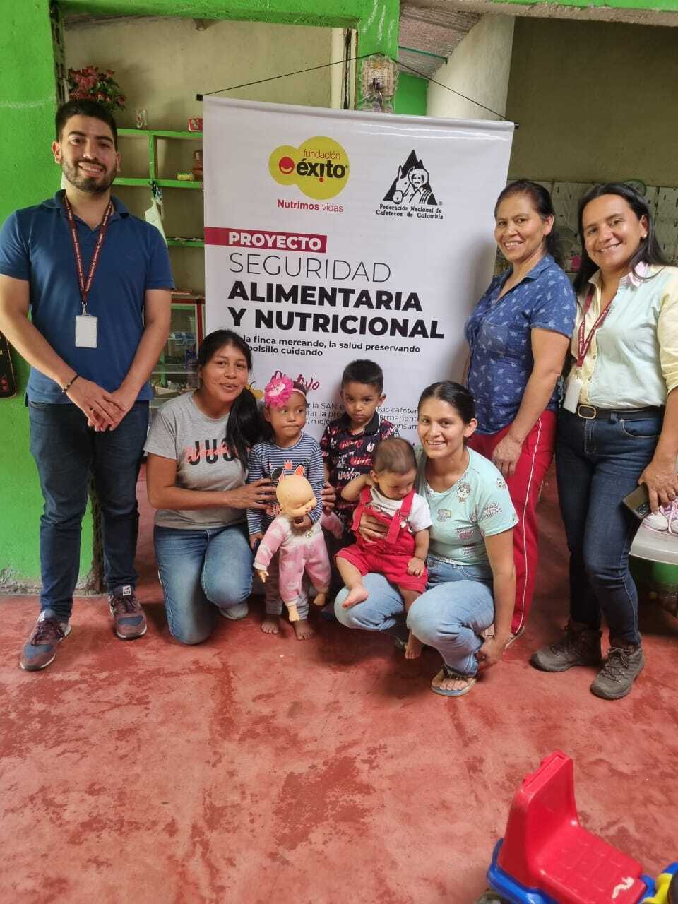 Grupo de adultos y niños pequeños posan para una foto frente a un cartel que anuncia un proyecto de seguridad alimentaria y nutricional. Los adultos y los niños podrían ser beneficiarios. La imagen tiene lugar en lo que parece ser un entorno comunitario o centro de atención infantil.